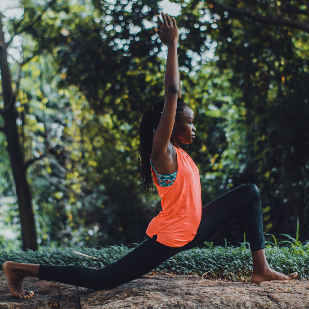 Woman in coral tank top and black yoga pants does yoga in the forest.